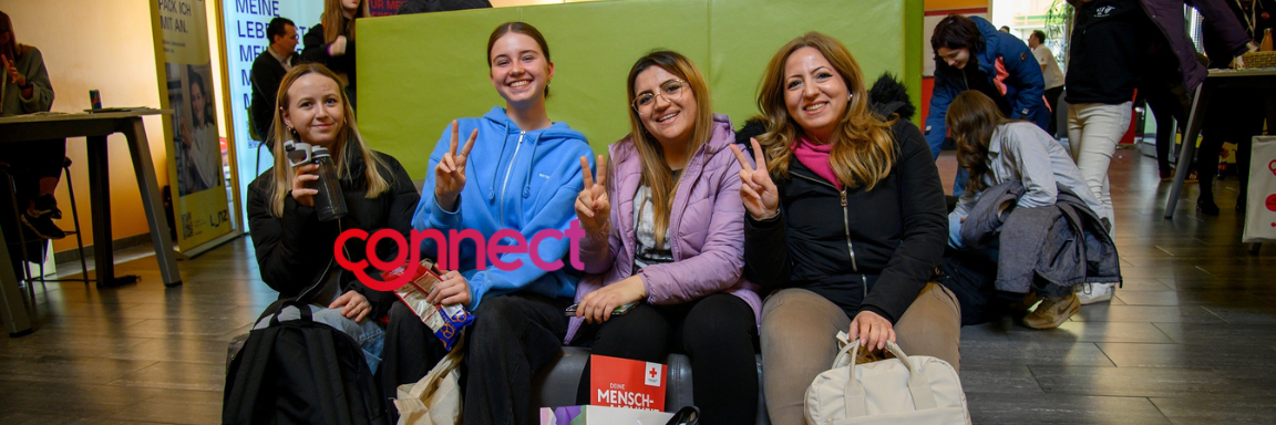 Junge Frauen sitzen auf einer Bank, lächeln und zeigen das Victory-Zeichen.