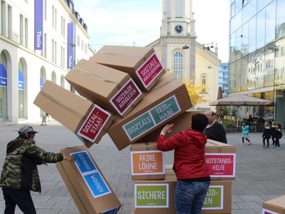 Es ist ein übergroßes Jenga aufgebaut, auf den Steinen stehen sozialstaatliche Begriffe. Hier stürzt das Jenga ein.
