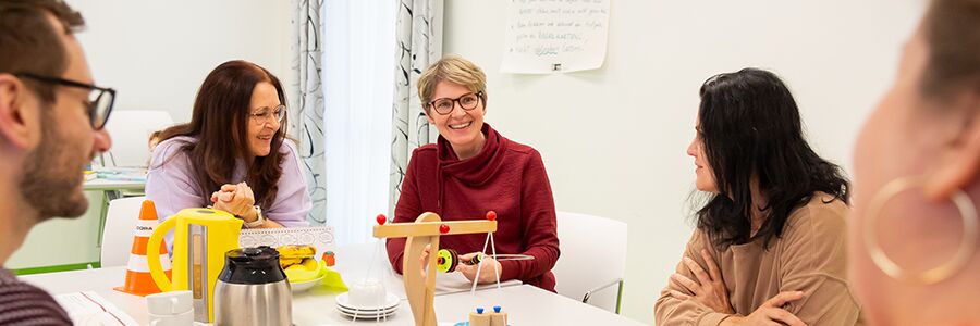 Menschen sitzen an einem Tisch mit Kaffeegeschirr und Spielzeug und tauschen sich aus.